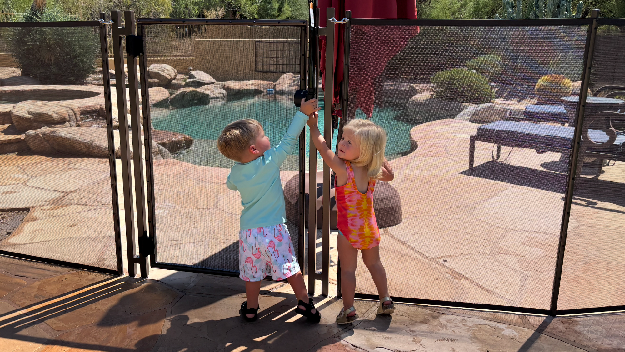 Two children interacting with a pool safety gate in a backyard setting.